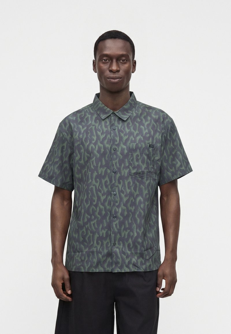 Homme portant une chemise à manches courtes avec un motif abstrait vert et foncé et un pantalon noir, debout devant un fond uni.