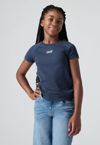Meisje met lange vlechten in een marineblauwe Levi's T-shirt en lichtblauwe jeans, staande met één hand op haar heup tegen een effen achtergrond.