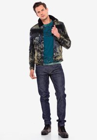 Schwarze und gelbe gemusterte Jeansjacke mit einem Lammfellkragen, blauem T-Shirt, dunkler Jeans und braunen Schnürstiefeln.
