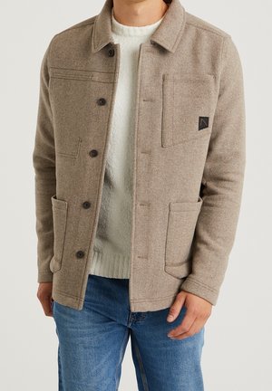 Man wearing beige buttoned jacket with chest and side pockets over white sweater and blue jeans, standing against plain background.