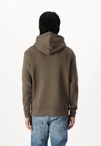 Brun hættejakke med snoretræk, lavet af bomuldsmateriale, ribbede manchetter og kant, båret sammen med lyseblå jeans. Enkel design uden synlige mønstre.