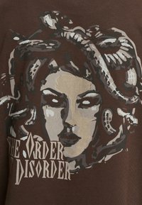 Brun t-shirt med et grafisk design af en kvindes ansigt med slanger, accentueret med nuancer af grå og beige. Teksten lyder "ORDEN AF UORDEN."