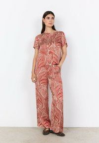 Kvinna iklädd matchande lös topp och vida byxor med abstrakt mönster i rött och beige samt svarta spikade ballerina-skor, stående mot en vit vägg.