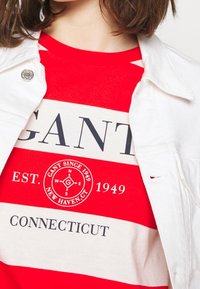 Punavalkoraidallinen T-paita, jossa "GANT"-logo, "EST. 1949" ja "CONNECTICUT"-tekstit; päällä valkoinen denimtakki.