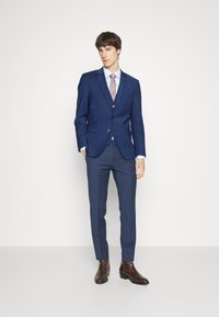 Costum de bărbați de culoare albastră, cu o croială ajustată, cu două nasturi, purtat cu o cămașă de culoare albastră deschisă și o cravată în dungi. Botine din piele texturată de culoare marron completează look-ul.