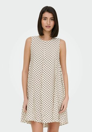 Mujer con cabello liso y oscuro que lleva vestido crema sin mangas con lunares negros, de pie contra un fondo claro y liso.