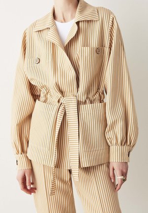 Beżowa i biała kurtka w pionowe paski, z przednimi kieszeniami, detalami z guzikami oraz pasem w talii, noszona z dopasowanymi spodniami.
