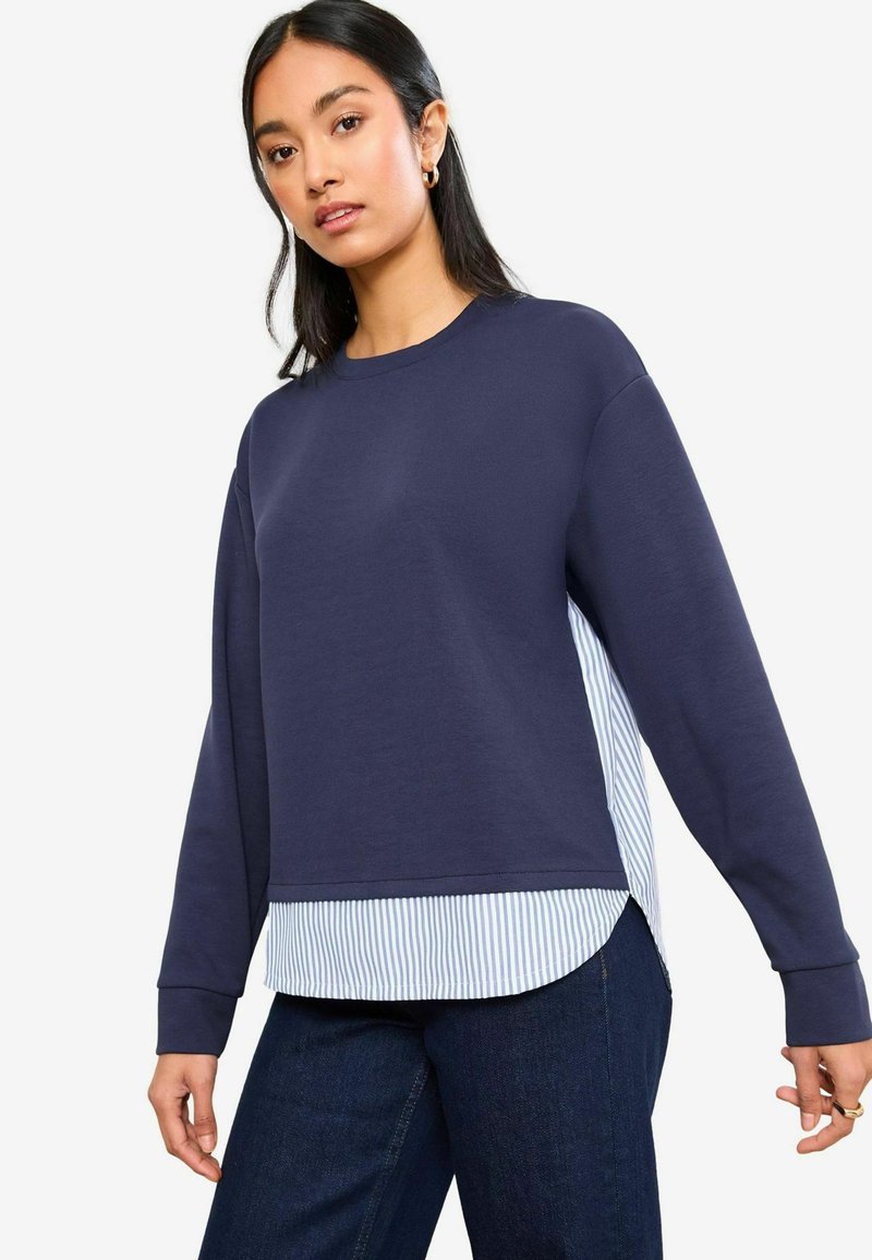 Felpa navy con maniche lunghe, sovrapposta a una camicia a righe leggera blu e bianca. Fit rilassato, scollatura tonda, polsini a costine.