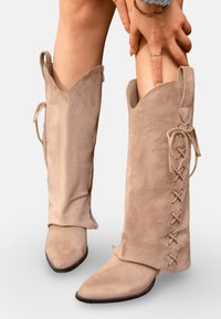 Bottes en suède beige arrivant jusqu'aux genoux avec un bout pointu, présentant des lacets sur le côté et un détail à nouer en haut, texture lisse et ajustement flexible.