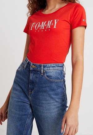 Vrouw draagt een rood Tommy Jeans t-shirt dat in een high waist blauwe spijkerbroek is gestopt, staat met ontspannen armen langs haar zij.