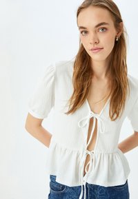 Top bianco a maniche corte con tessuto testurizzato, scollo a V profondo, dettaglio legato sul davanti e orlo peplum. Abbinato a jeans in denim blu.