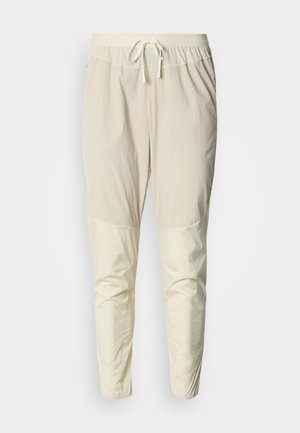Pantalon de jogging beige clair avec taille élastique, cordon de serrage et finition semi-brillante. Présente des panneaux genoux plus clairs distincts.