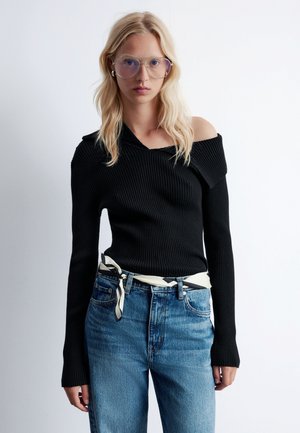 Black ribbed knit sweater with an asymmetrical neckline, paired with high-waisted blue denim jeans and a patterned white and black belt.