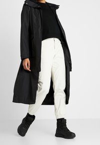 Long manteau imperméable noir par-dessus un pull noir, pantalon blanc fuselé et bottes noires à lacets, avec capuche et poches latérales.