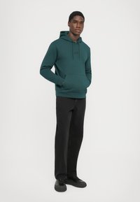 Felpa verde acqua con tasca a marsupio e cappuccio regolabile; presenta un logo sottile in nero. Abbinata a pantaloni neri e sneakers scure.