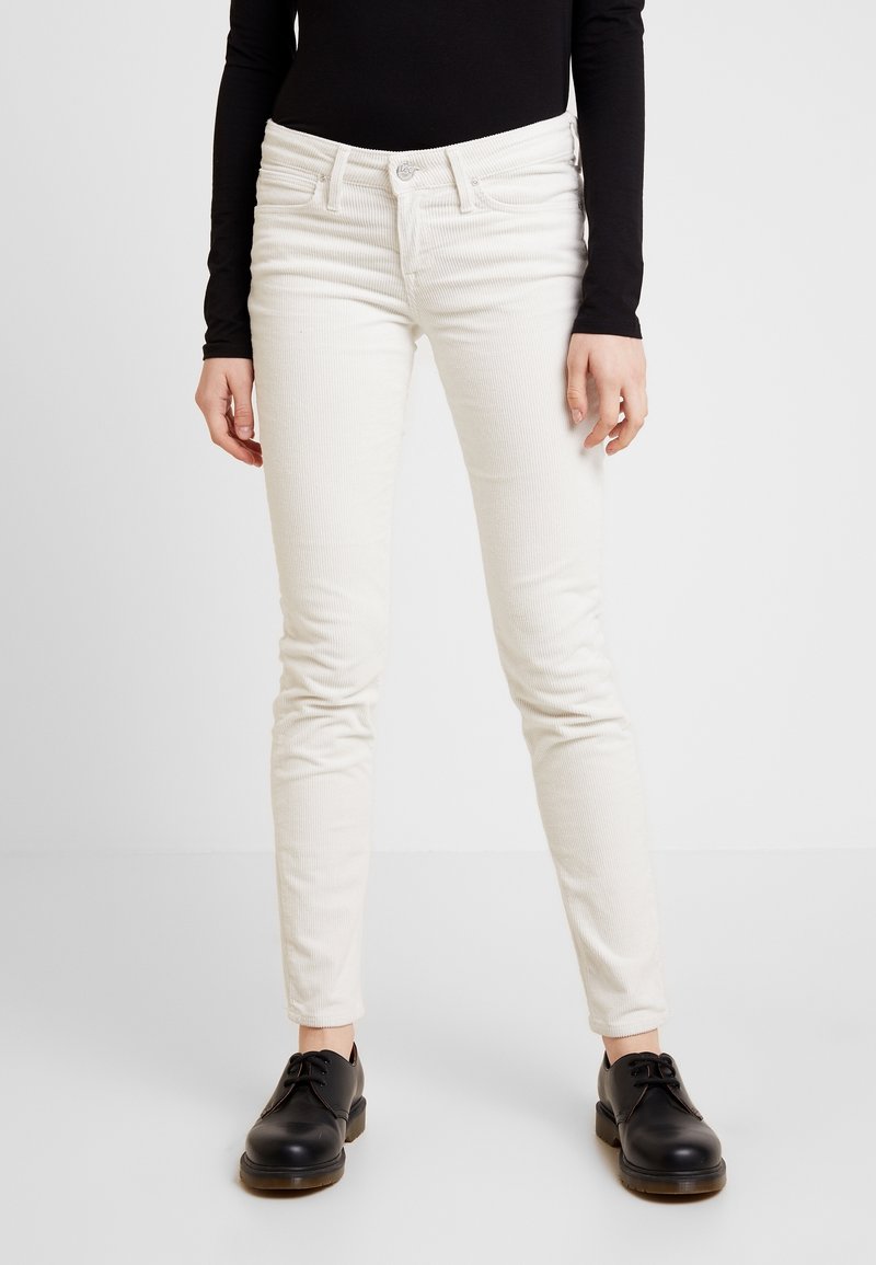 Person som bär vita kordbyxor, svart långärmad topp och svarta snörskor som står på vit bakgrund.