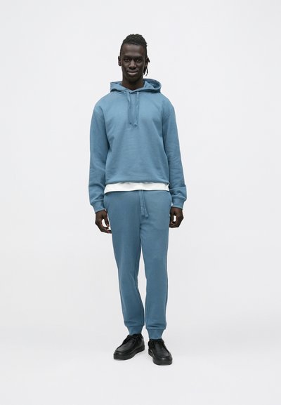 Hoodie bleu clair avec cordon de serrage et pantalon de survêtement assorti, tous deux en tissu doux, associés à des baskets noires et un t-shirt blanc en dessous.