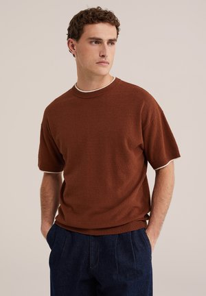 Jeune homme aux cheveux bouclés portant un pull marron à manches courtes et un pantalon bleu foncé, debout les mains dans les poches, regardant vers la droite.