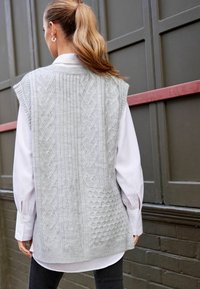 Gilet en tricot gris clair avec divers motifs texturés, manches courtes et col côtelé, porté par-dessus une chemise blanche à col.