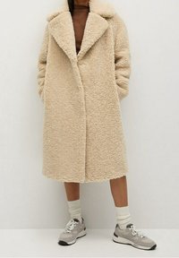 Beige keinanan turkki leveällä kauluksella, etupainikkeilla ja sivutaskuilla, yhdistettynä harmaisiin lenkkareihin ja valkoisiin sukkiin.