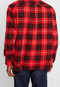Camisa de franela de cuadros rojos y negros con mangas largas, que presenta un corte holgado y puños abotonados. La textura parece suave y ligeramente cepillada.