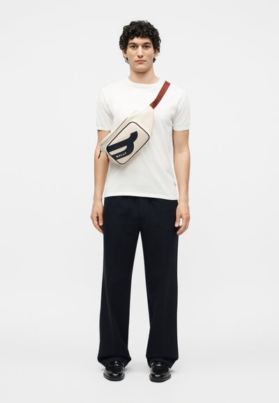 Άνδρας που φοράει λευκό μπλουζάκι, μαύρο παντελόνι με φαρδιά μπατζάκια, μαύρα παπούτσια και μπεζ τσάντα ώμου με μαύρες και καφέ λεπτομέρειες.