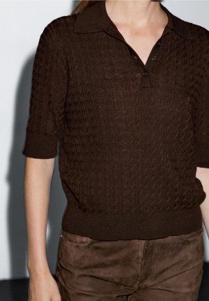 Femme portant un haut en maille texturée marron foncé avec des manches courtes et un col, associé à un pantalon en daim marron.