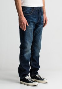 Donkerblauwe spijkerbroek met een klassieke rechte snit, met een versleten textuur en contrasterende stiksels, gedragen met zwarte canvas sneakers.