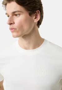 Biały bawełniany t-shirt z okrągłym dekoltem i małym, wytłaczanym logo na piersi. Gładka faktura, krótkie rękawy, dopasowany krój.