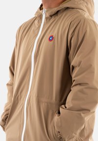 Giacca a vento marrone chiaro con cappuccio realizzata in tessuto morbido, caratterizzata da una cerniera bianca, polsini elasticizzati e un piccolo logo circolare sul lato sinistro del petto.