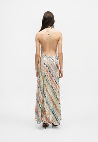 Mujer con un vestido largo de tirantes con rayas onduladas multicolores y espalda descubierta, de pie de espaldas sobre un fondo blanco liso.