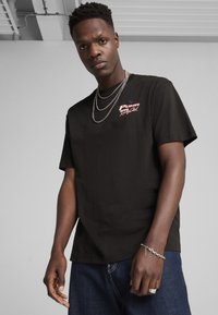 Sort bomulds t-shirt med korte ærmer, med rød "Puma" logo, rund halsudskæring og lagede sølv kæder. Denim jeans synlige nedenfor.