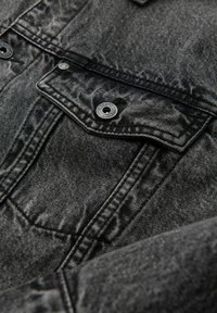 Giubbotto in denim nero con tessuto strutturato, dettagli con bottoni a scatto e una tasca frontale. Caratterizzato da un design classico con cuciture visibili.