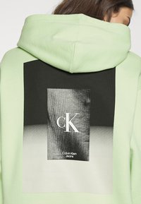 Persoon draagt een lichtgroene hoodie met een zwart-wit rechthoekig Calvin Klein Jeans-logo op de achterkant, met de initialen cK.