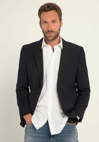 Marineblazer met zwarte revers, witte overhemd eronder en blauwe spijkerbroek. Heeft een enkele knoopsluiting en een borstzak.
