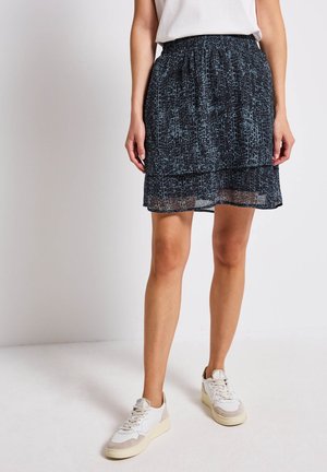 Zwarte rok tot de knie met een geprofileerd patroon en een doorzichtige overlay, voorzien van een elastische tailleband. Gedirigeerd met witte en beige sneakers.