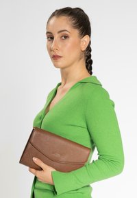 Bolso de mano de cuero marrón con un acabado Texturizado y cierre de solapa, sostenido contra una blusa verde de canalé.