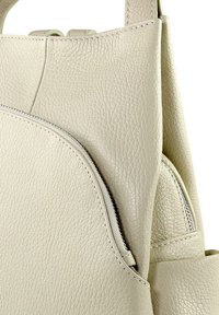 Sac en cuir gris clair avec une surface texturée, une poche ronde zippée et des bretelles plates. Le design met l'accent sur la simplicité et la fonctionnalité.