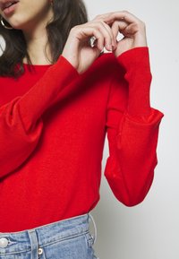 Roter Pullover mit langen Ärmeln und gerippten Bündchen, der eine glatte Textur aufweist. Kombiniert mit hochgeschnittenen hellen Denim-Jeans.