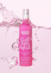 Flacon pulvérisateur transparent contenant un liquide rose. L'étiquette mentionne "Umberto Giannini Curl Jelly Spray Revitalisant pour Boucles", 150 ml, avec des éclaboussures d'eau.