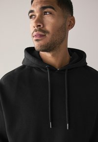 Sudadera negra con capucha y cordón, con una textura suave y un ajuste holgado. Vista en primer plano que muestra el rostro y el cuello de un joven.