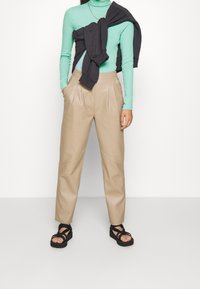 Hellgrüner Rollkragenpullover, beigefarbene, plissierte Hose und schwarze Sandalen mit Riemchen. Das Oberteil hat lange Ärmel; das Outfit umfasst einen schwarzen Pullover, der um die Taille gebunden ist.