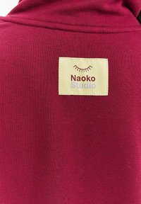 Bordowy materiał tekstylny o miękkiej, gładkiej fakturze z prostokątną kremową metką. Na metce widnieje napis „Naoko Studio” w fioletowo-czerwonych czcionkach.