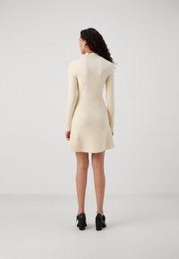 Abito a maniche lunghe di maglia color crema con corpetto aderente e gonna svasata, caratterizzato da un collo alto e una texture morbida. Scarpe con tacco nere completano il look.
