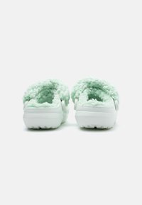 Une paire de sabots blancs à enfiler avec une doublure en molleton duveteux vert menthe, présentée de dos sur un fond blanc uni.