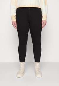 Calças de ganga pretas justas com uma textura suave, apresentando um fecho de botão frontal e botões decorativos laterais, combinadas com botas bege de atacadores.