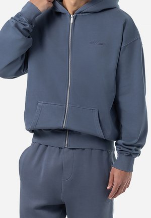 Mężczyzna w niebieskiej bluzie z kapturem zapinanej na zamek i dopasowanych spodniach dresowych, ręką poprawiający kaptur, jednolite tło.