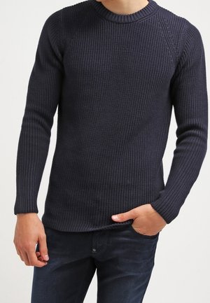 Mand iført en mørk marineblå ribstrikket rundhalset sweater med lange ærmer og mørke jeans, hånd i lommen mod en ensfarvet lys baggrund.