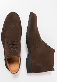 Bottines en cuir suédé marron à lacets avec détails brogue et semelles en caoutchouc noir, présentées sur un fond blanc.