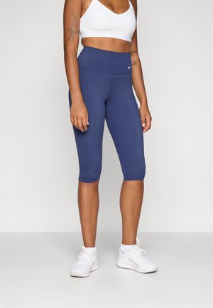 Navy blue capri leggings with a high waistband, made of stretchy fabric, paired with a white sports bra and white athletic shoes.